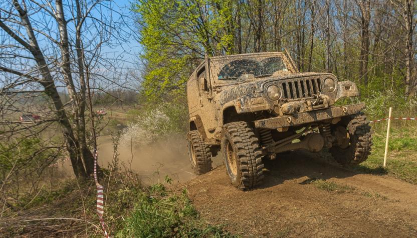 Ein schlammiger Geländewagen fährt über eine unbefestigte, staubige Strecke im Wald, umgeben von Absperrband und Bäumen.