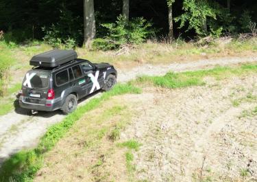 Schwarzer Geländewagen mit Dachbox und weißem X  auf Beifahrertür fährt einen Feldweg entlang. Links befindet sich ein Wald und rechts eine Wiese 