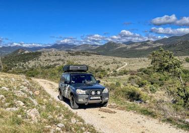 Ein schwarzer Geländewagen mit Dachbox fährt auf einem steinigen Pfad durch eine hügelige, trockene Landschaft. Im Hintergrund erstrecken sich Berge unter einem blauen Himmel mit einigen Wolken.
